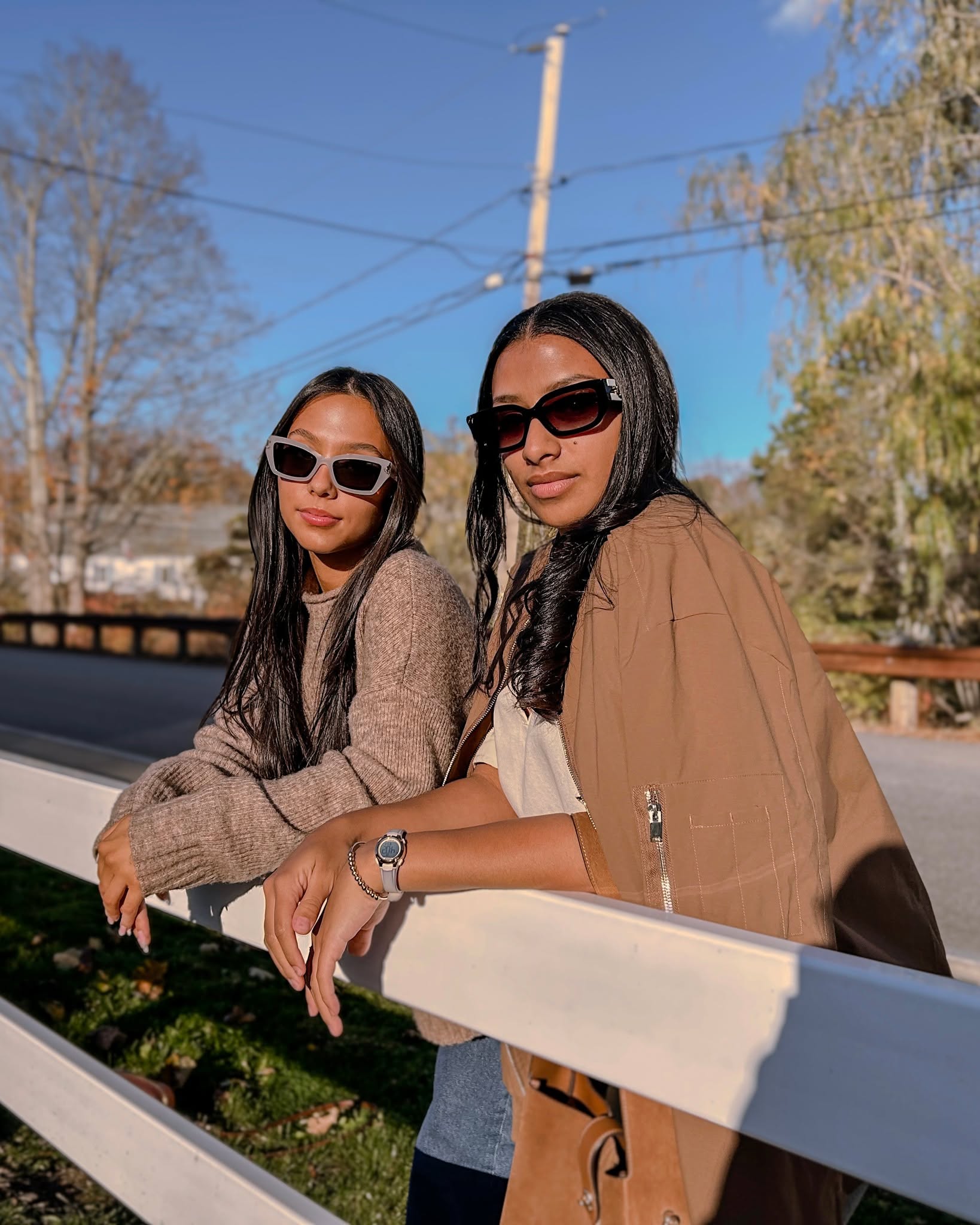 Two women wearing trendy sunglasses, one in light-colored frames and the other in dark frames, leaning against a white fence, showcasing fashionable eyewear in an outdoor setting.